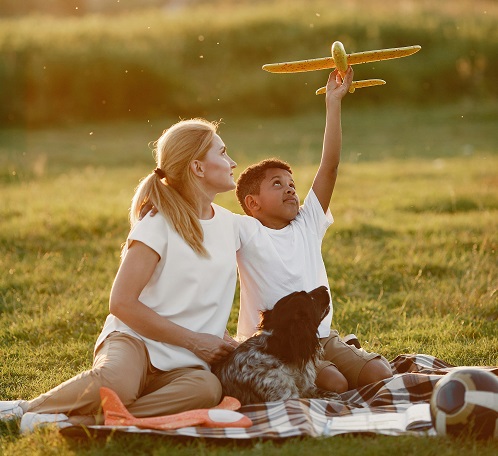 Vrouw met kind en hond zit op de grond op een picknickkleed. Het jongetje heeft een vliegtuig in z'n hand.